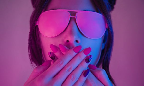 Retrowave style portrait of young woman in pink glasses. Young woman in pink glasses covers her mouth with hands with a manicure, retrowave style portrait. Demonstration of a beautiful manicure.